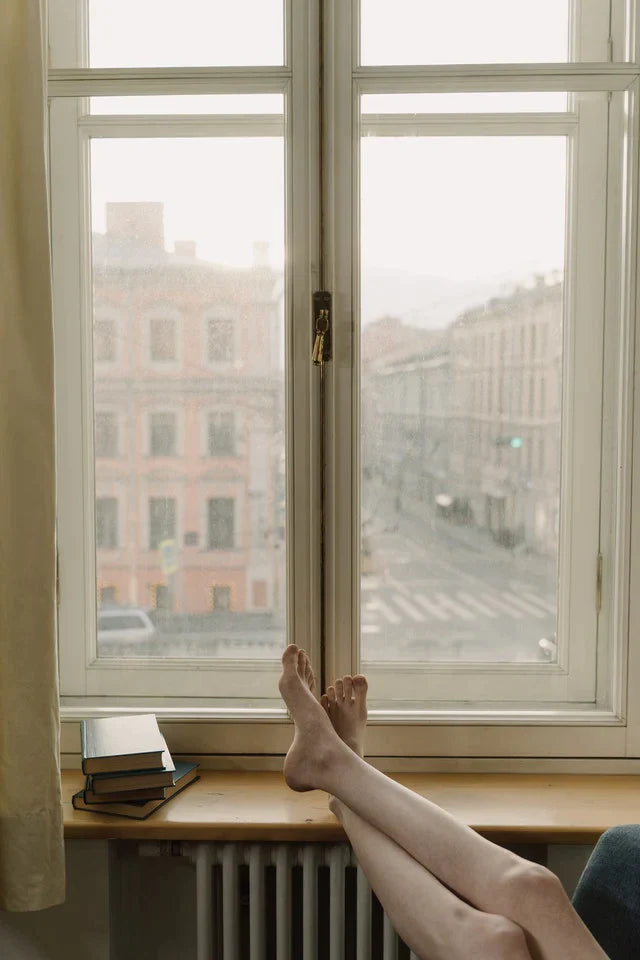Junge Frau mit Sonnenbrille sitzt an ihrem Fensterbrett bei geöffnetem Fenster und trinkt ihren Kaffee am Morgen