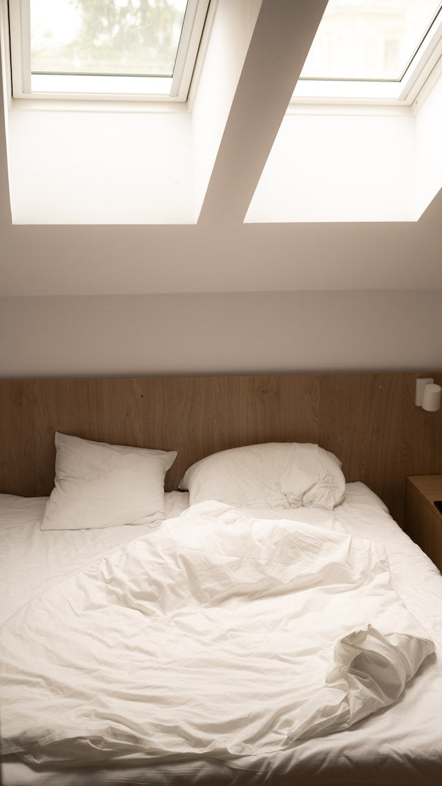 Gesunder Schlaf: Blick in ein Schlafzimmer. Man sieht ein Holzbett mit weißer Bettwäsche darüber zwei große Fenster an einer Dachschräge