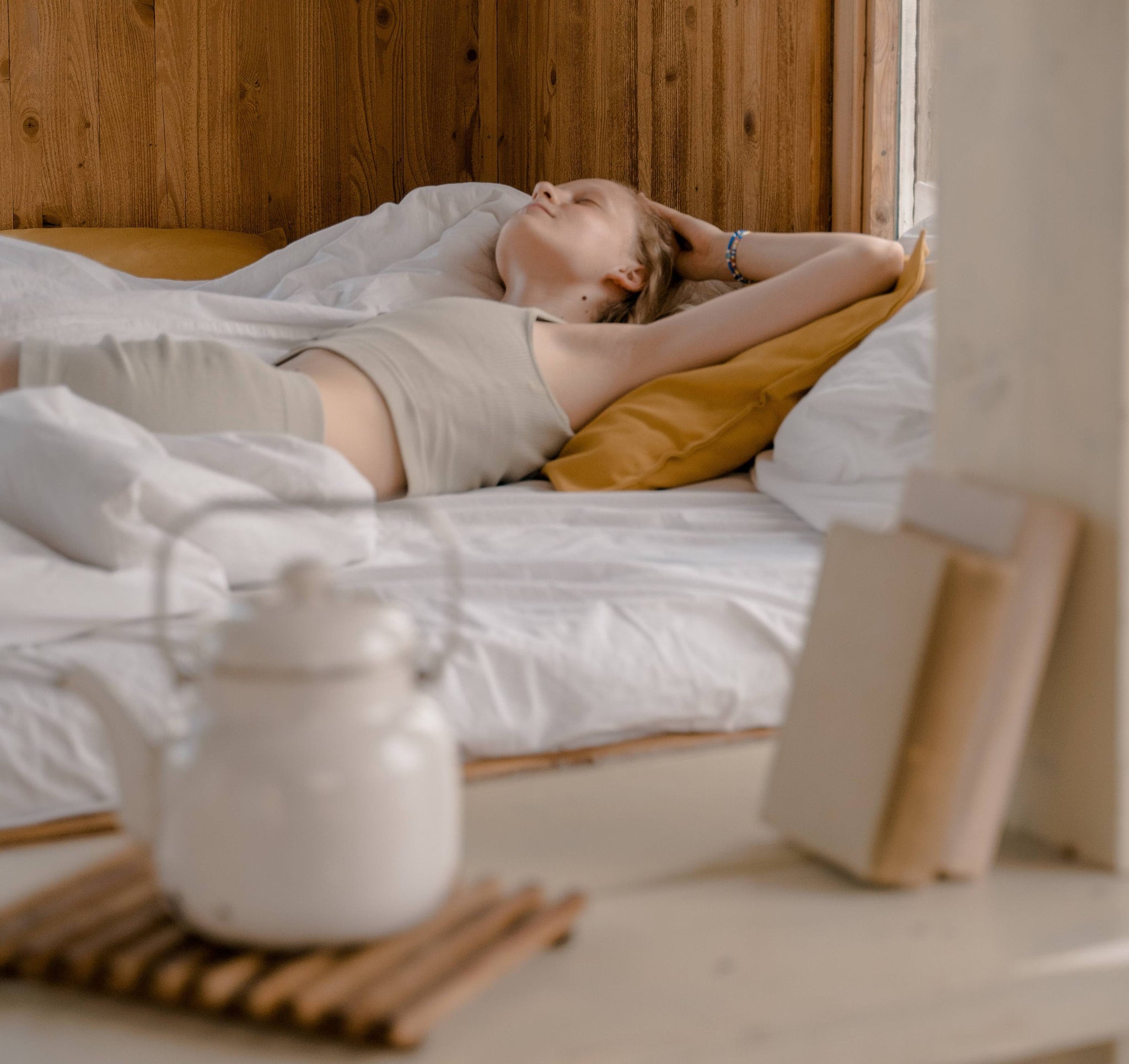 Frau im beigen Top und beiger Hose liegt mit geschlossenen Augen im Bett. Im Vordergrund ein Regal mit einer weißen Teekanne und zwei weißen Büchern.